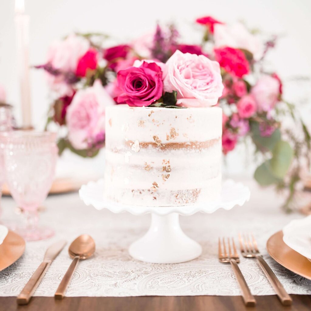 Elegant naked wedding cake decorated with pink and red roses, displayed on a white cake stand with floral table setting.