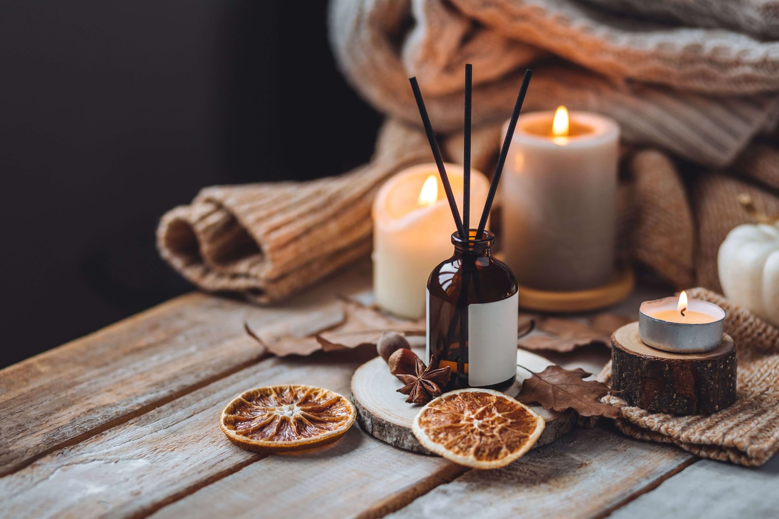 Cozy autumn flat lay with lit candles, amber glass reed diffuser, dried orange slices, star anise, a