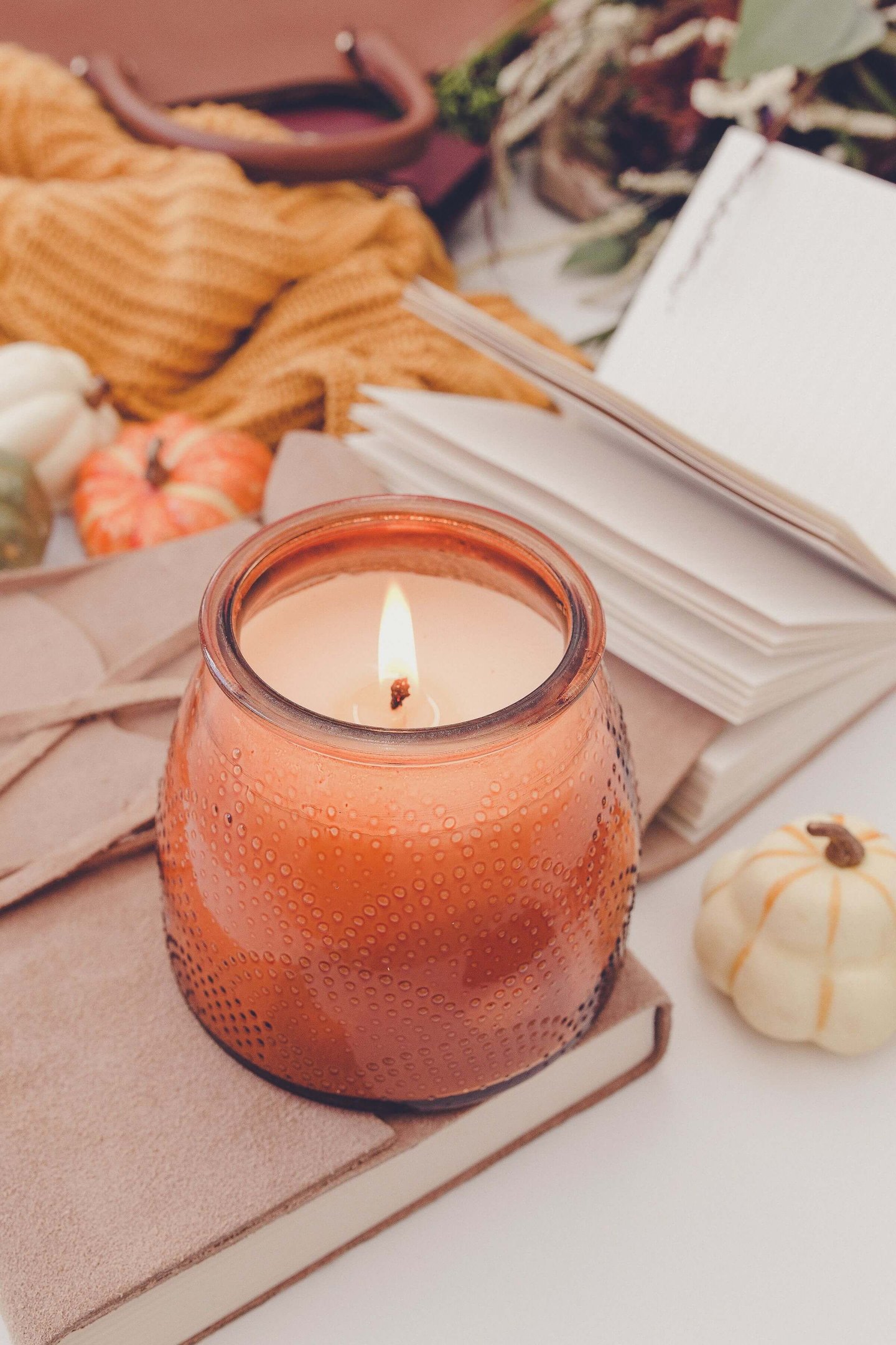 Lit orange candle in a textured glass jar, resting on suede notebooks with an open journal, mini pum