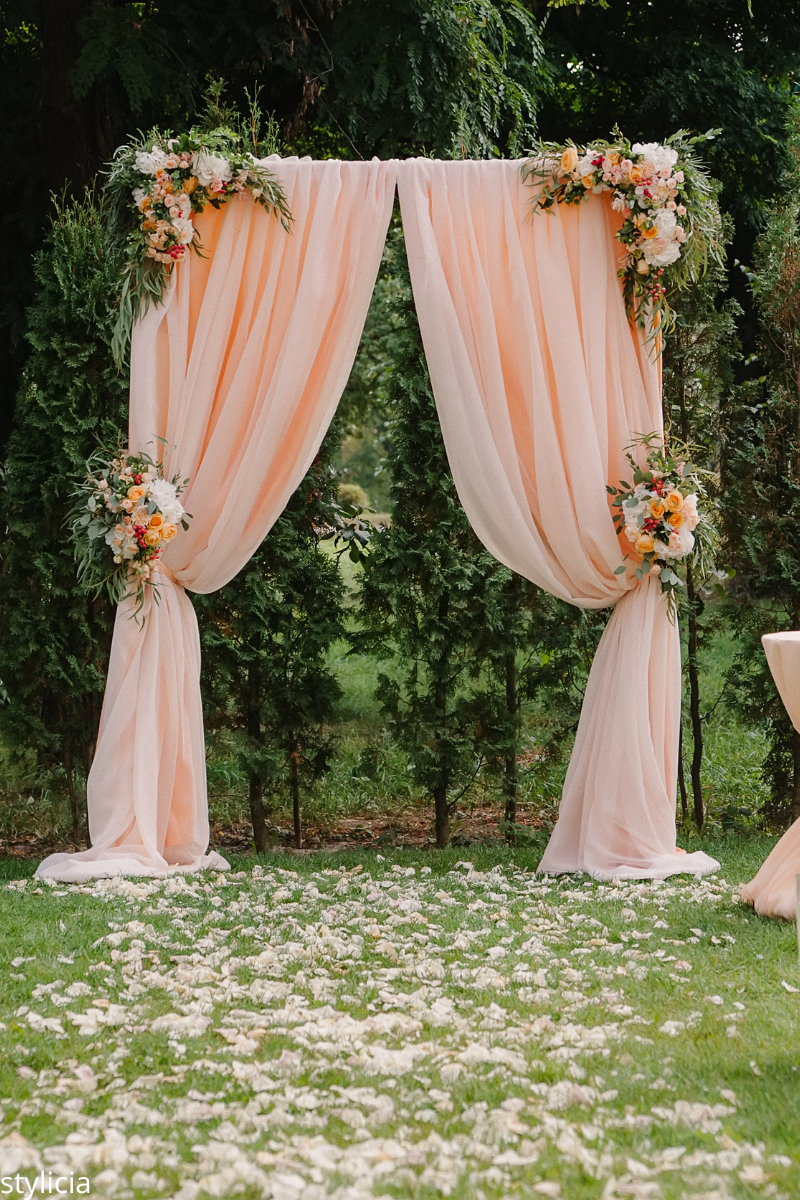 Outdoor wedding arch decorated with peach drapes and floral arrangements, showcasing a romantic wedding color trend in sunset hues.