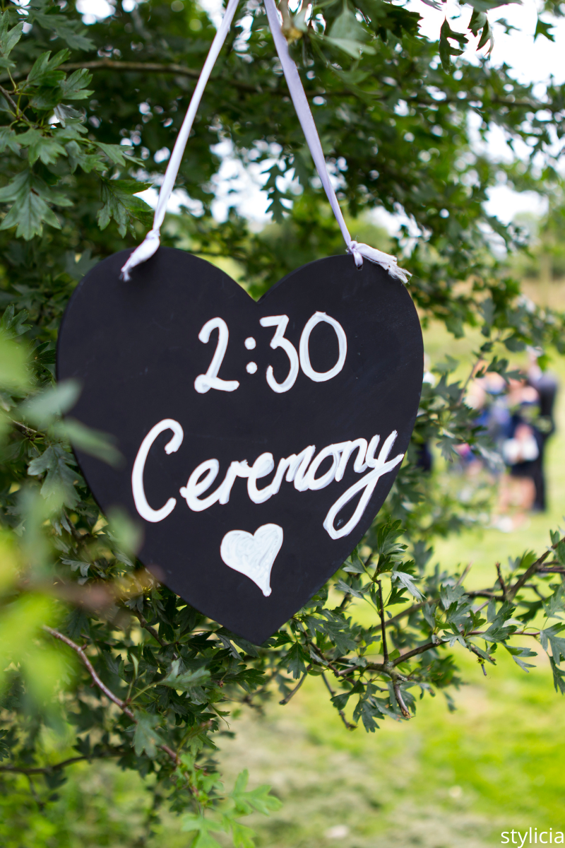 Black heart-shaped wedding sign with ceremony time written in white, representing things wedding guests don’t care about details.