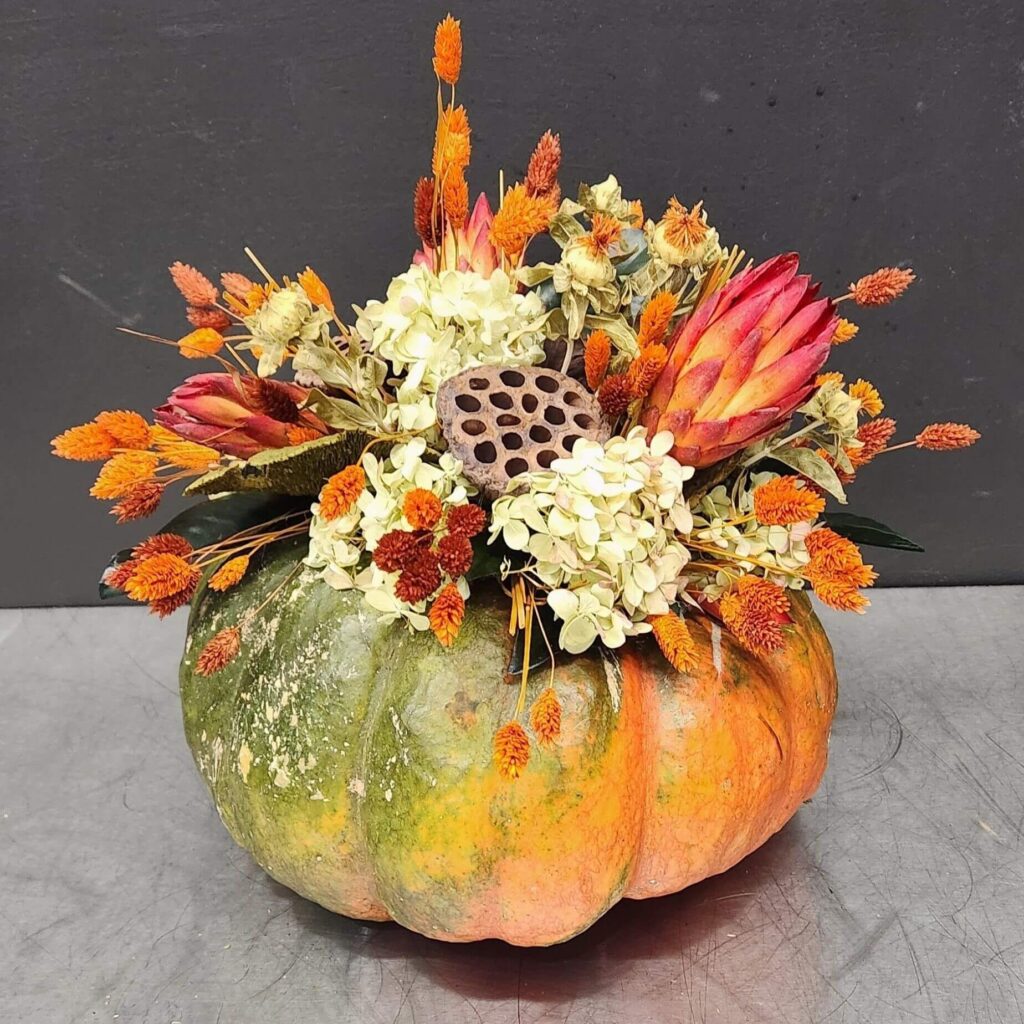 Rustic pumpkin centerpiece filled with dried autumn flowers, hydrangeas, protea, and vibrant orange accents – perfect Thanksgiving table decoration idea.