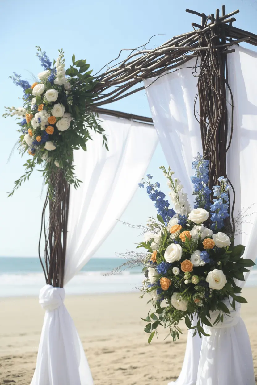 Bare branch wedding arch lashed into a frame on a beach, with white drapes and clusters of blue, white and peach blooms across the top corner