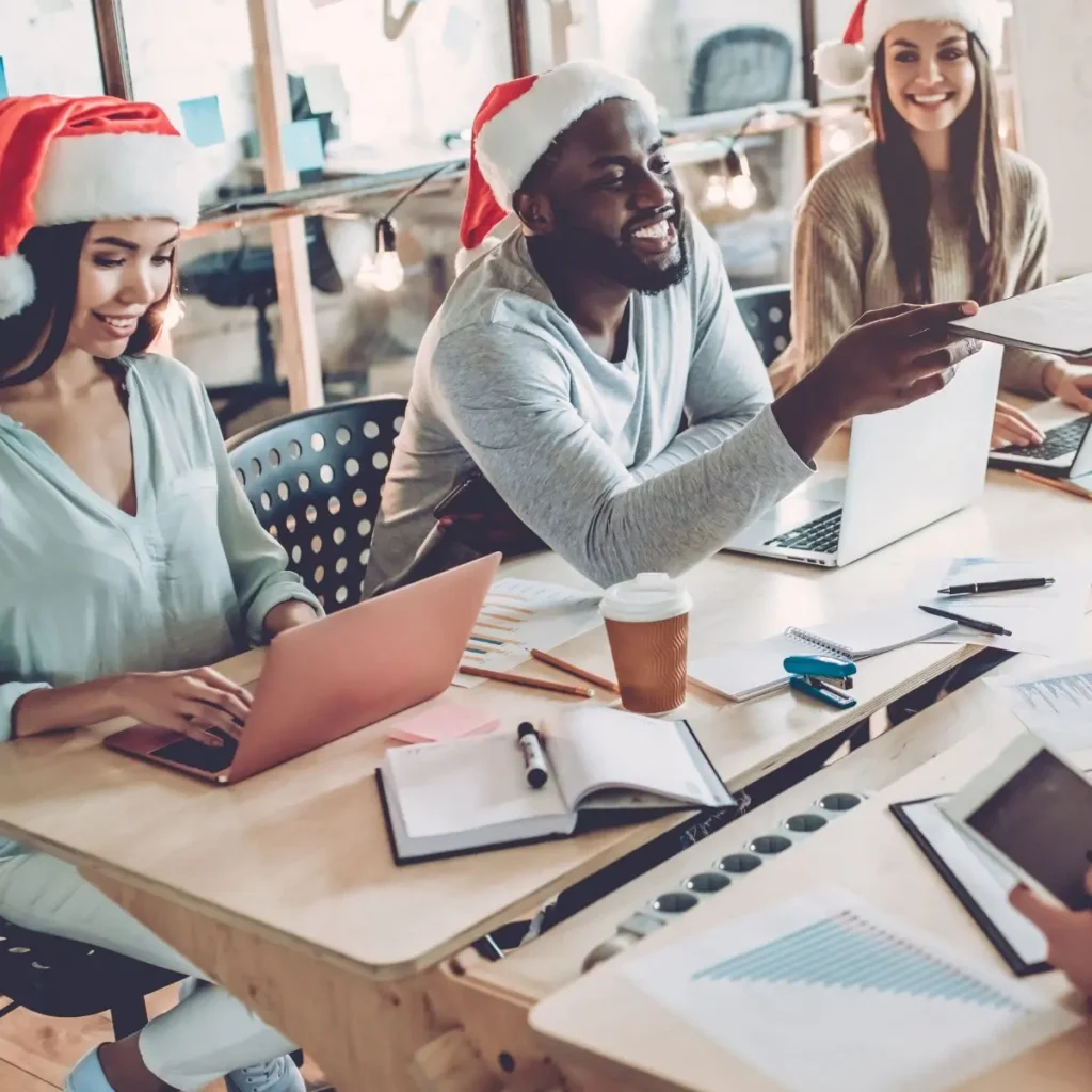 Coworkers wearing Santa hats working together in an office during a Christmas celebration, smiling and enjoying the holiday atmosphere.