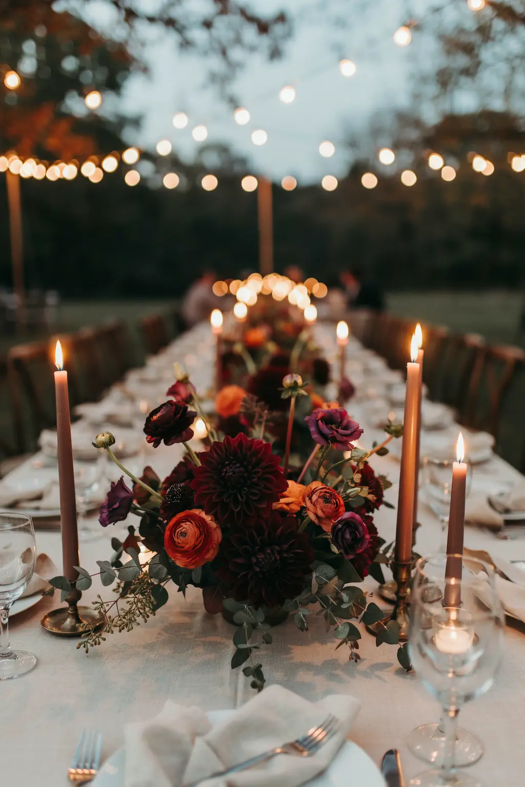 Fall wedding flowers under warm string lights, where burgundy dahlias read almost black by dinner