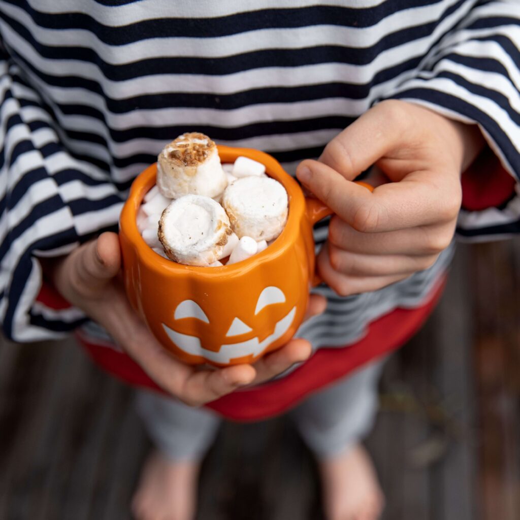 Child holding a pumpkin-shaped Halloween mug filled with hot chocolate, marshmallows, and toasted toppings – cozy festive drink idea.