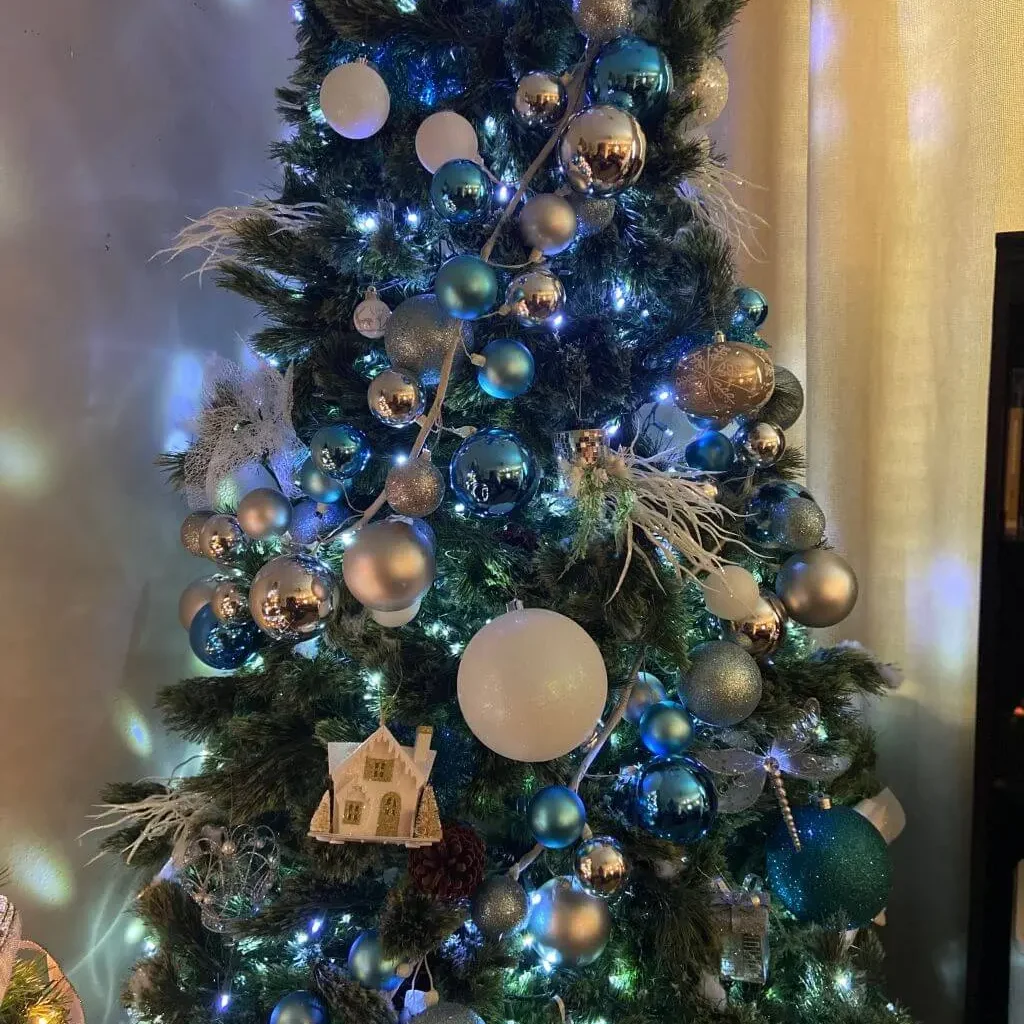 Blue and silver Christmas decoration on a green tree with frosted branches, shiny silver baubles, and soft lights creating a modern winter look.