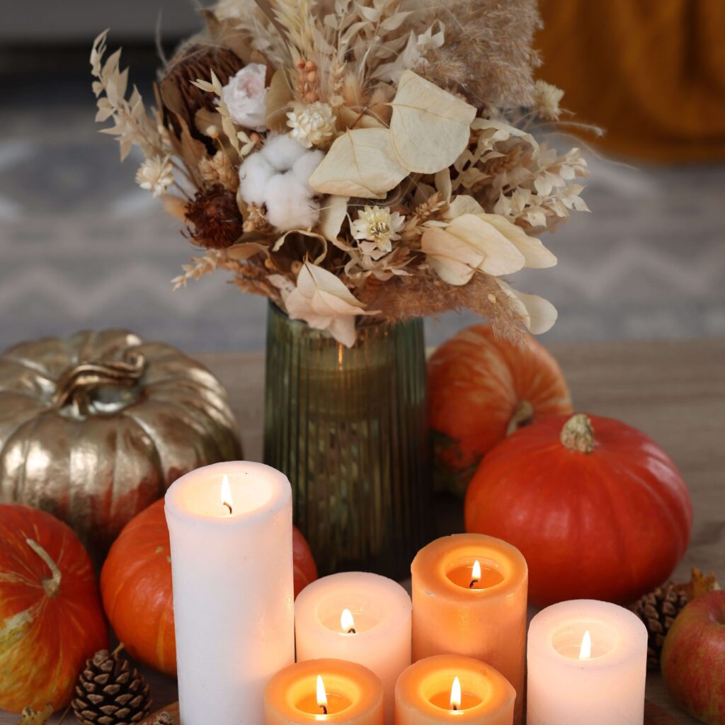 Cozy fall decoration with lit candles, pumpkins, and a dried flower bouquet in a green vase on a wooden table.