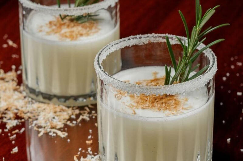 Overhead flat lay of snowy white Winter Wonderland Mocktail with rosemary garnish, surrounded by orange slices, white chocolate, and pinecones for Christmas décor.